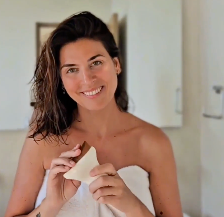 A smiling woman wrapped in a white towel holds our Lymphatic Drainage Brush in both hands, photographed indoors against a softly blurred light background.
