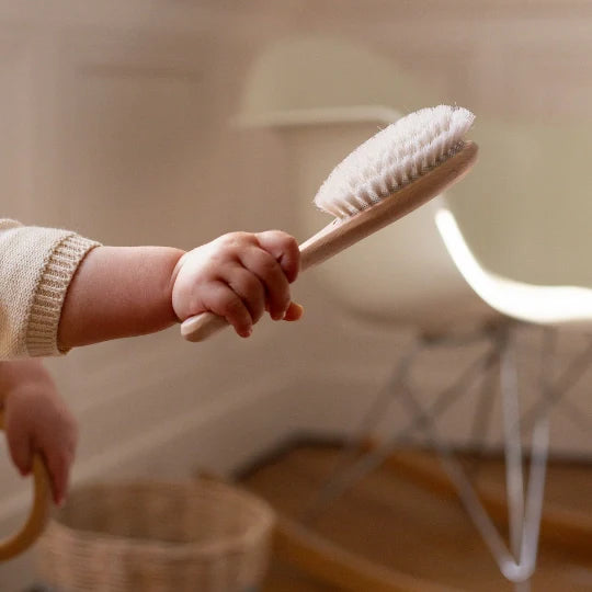 Baby hand holding a Bachca beechwood hair brush with ultra-soft bristles, in a warm interior with wooden floor and designer chair in the background.