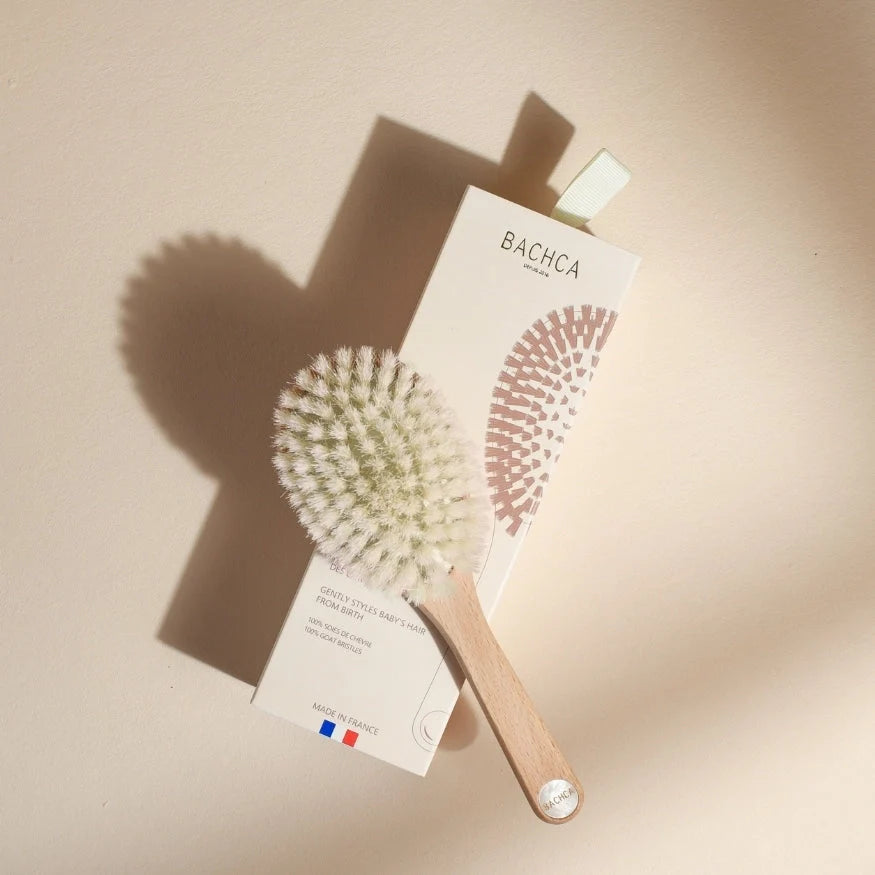 Bachca baby hair brush positioned diagonally on its cream case, with light wood handle and very soft white bristles, lit by natural light on a soft beige background.