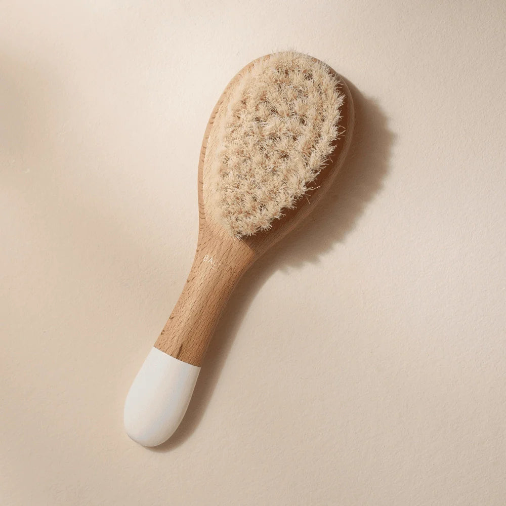 Small Bachca baby hair brush with soft goat hair bristles and a wooden handle, photographed on a pale pink background.