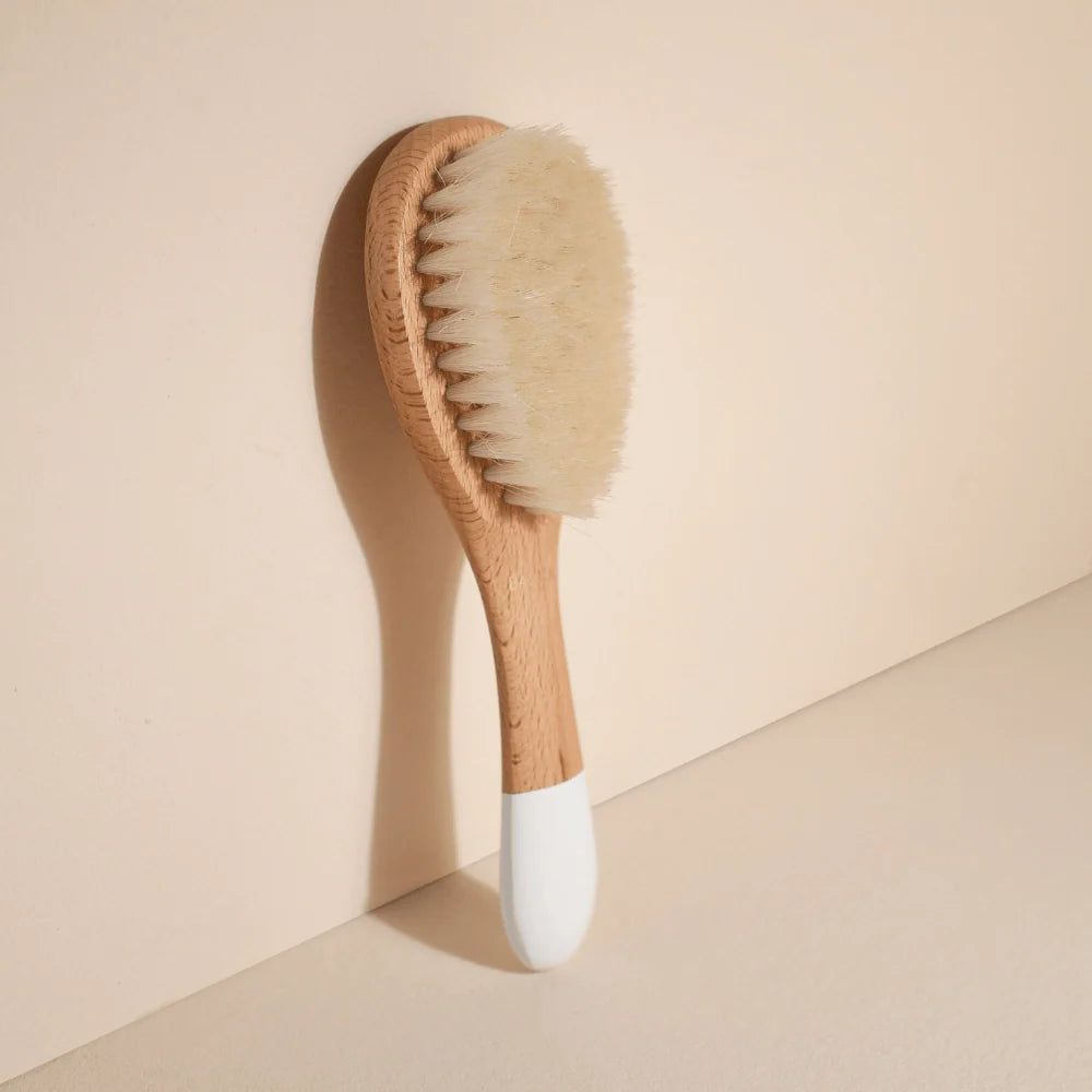Small Bachca baby hair brush with soft goat hair bristles and a wooden handle, photographed leaning against a pale pink background.