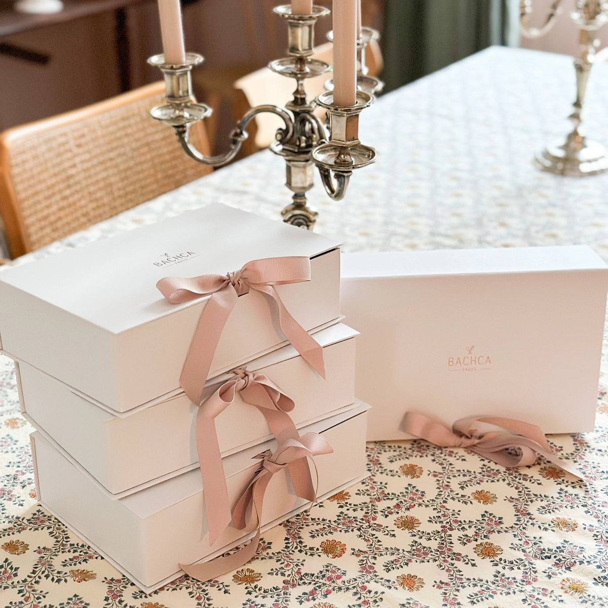 Stack of white boxes with pink ribbons on a floral tablecloth, with a candelabra in the background.
