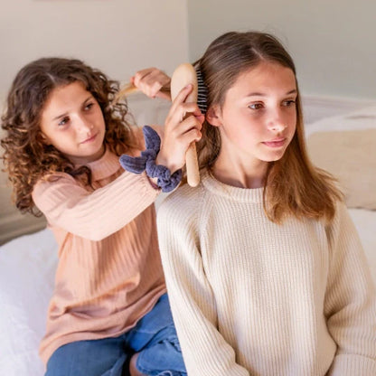 Little girl styling another child’s hair with a Bachca wood Everyday Detangling Hair Brush, sitting on a bed in a bright room.