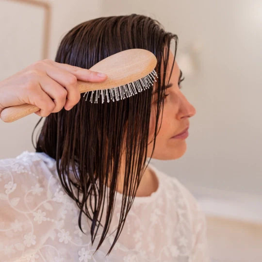 Woman styling her wet hair with a Bachca Everyday Detangling Hair Brush in wood, in a bright bathroom, wearing a white lace top.