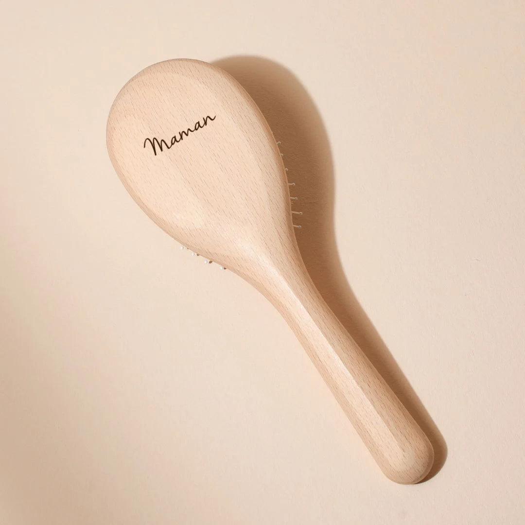 Bachca hairbrush in natural beechwood, back view, with the word “Maman” delicately engraved on the handle, placed on a powder beige background.