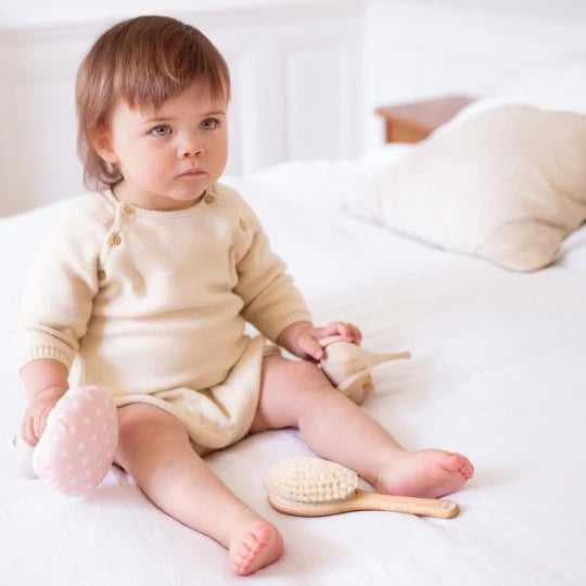 Baby sitting on a white bed, wearing a beige onesie, holding a toy and a wooden spinning top, with a Bachca beechwood hair brush with ultra-soft bristles placed near their feet.