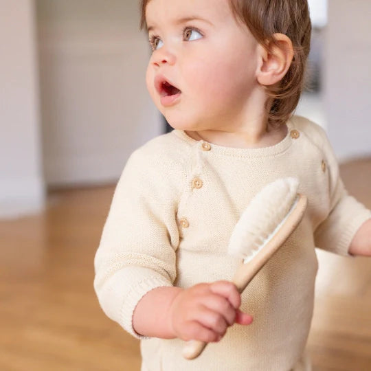 Small child with light eyes wearing a beige onesie, holding a Bachca beechwood hair brush with soft bristles, in a bright interior with wooden floor.