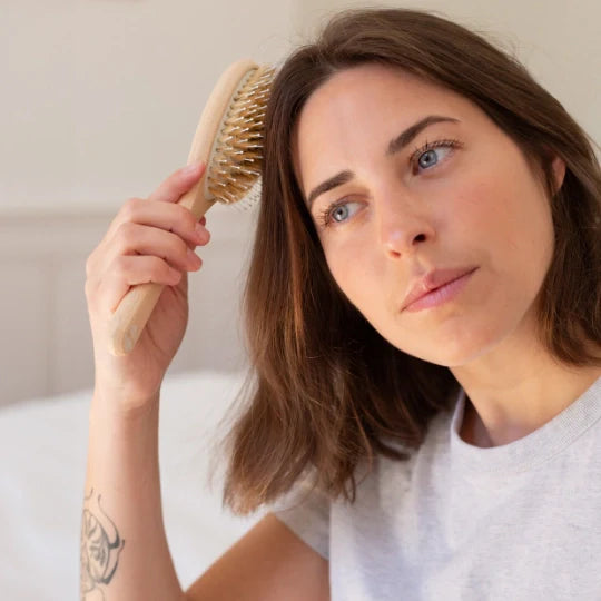 Woman brushing her hair with a natural beechwood Bachca hair brush with nylon and boar bristles, seated in a bright room wearing a light gray t-shirt.