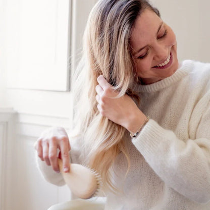 Smiling woman brushing her hair with a natural wood Bachca hair brush, wearing a white sweater in a bright interior.