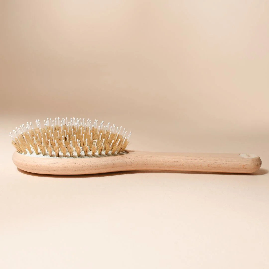 Bachca hair brush laid flat on a beige background, side view. Its natural beechwood handle extends into a cream cushion with nylon and boar bristles, finished with a pearlescent medallion at the end.