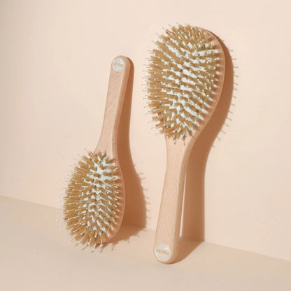 Two Bachca hair brushes hanging against a beige wall: on the left, the small size, on the right, the large size. Both feature a beechwood handle, nylon pins and boar bristles, with a pearlescent medallion at the end of the handle.