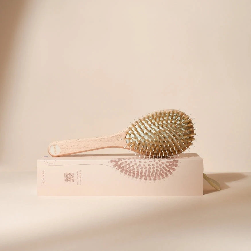 Bachca hair brush photographed from the side, placed on its illustrated box, with a light wood handle and cushion filled with mixed bristles, on a soft beige background.