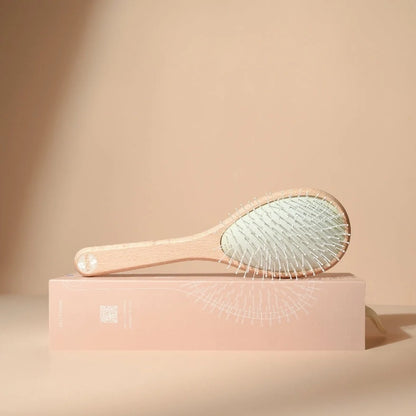 Bachca Everyday Detangling Hair Brush, side view, placed on its illustrated blush pink box, with a light beechwood handle and cushion with fine bristles, on a neutral beige background.