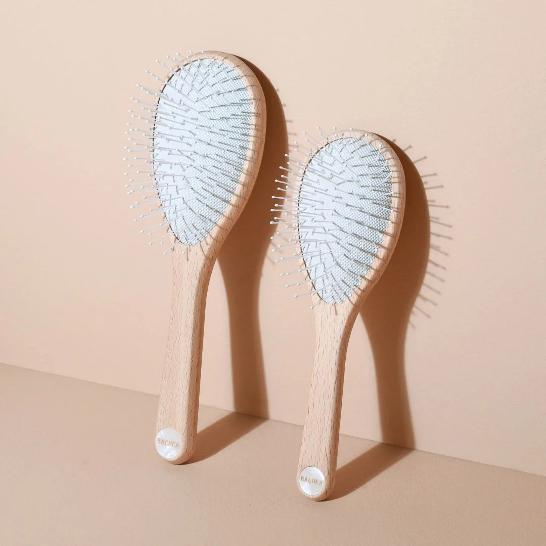Two Bachca Everyday Detangling Hair Brushes, large and small sizes, in raw beechwood with white nylon bristles, standing against a beige wall.