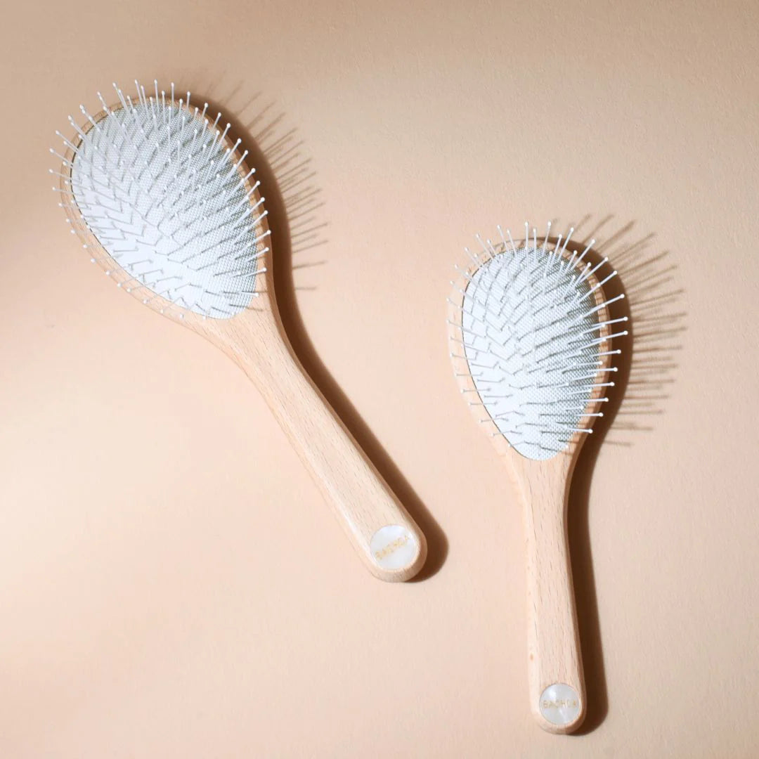 Two Bachca Everyday Detangling Hair Brushes, small and large sizes, in raw beechwood with white nylon bristles, laid flat on a beige background.