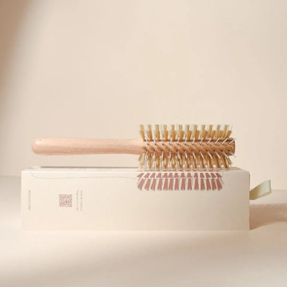 Bachca round styling brush in light wood, laid horizontally on its cream box, side view emphasizing the white and beige mixed bristles and illustrated packaging.