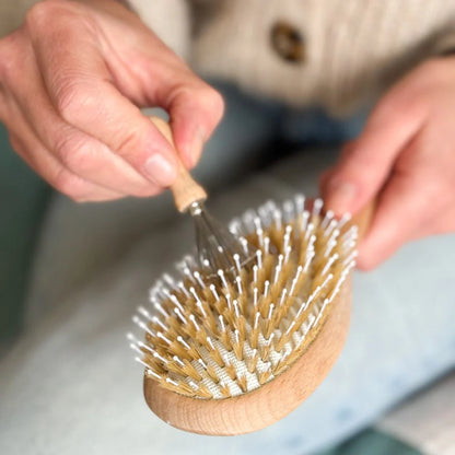 Hand using a Bachca wooden hairbrush with a metal comb to remove hair and residue.