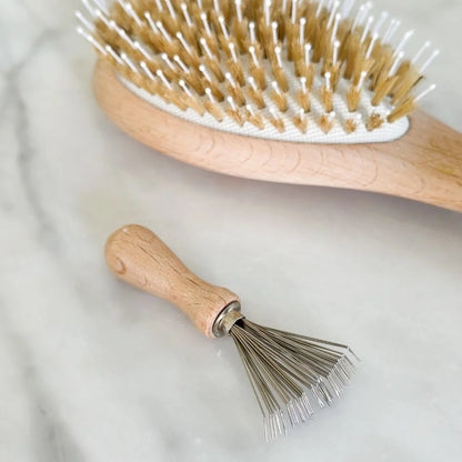 Bachca hairbrush cleaning comb in wood, positioned on marble next to a boar bristle brush.