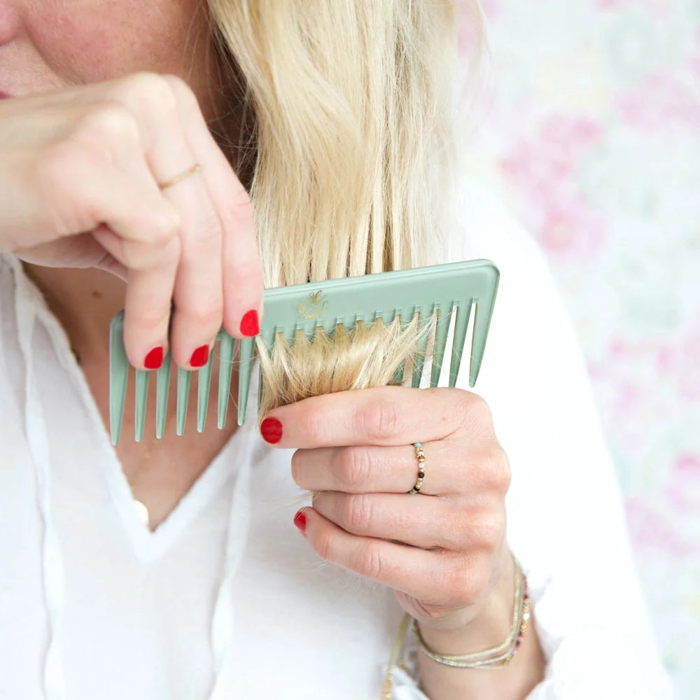 Woman styling her blonde hair with the Bachca Opaline green comb, wearing a white outfit and red nail polish, set against a soft pastel floral background.