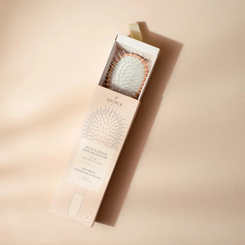 Young girl with curly hair brushing her lengths with the Bachca detangling and volume brush in light wood, wearing a pale pink top.