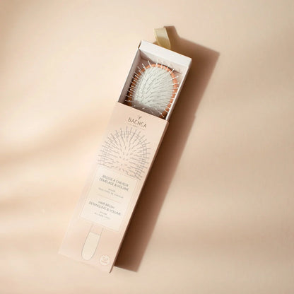 Young girl with curly hair brushing her lengths with the Bachca detangling and volume brush in light wood, wearing a pale pink top.