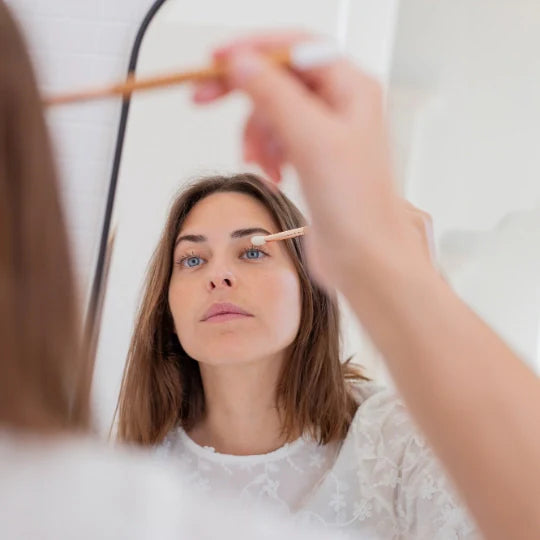Woman applying eyeshadow in front of a mirror using the Bachca angled eyeshadow brush, featuring a light wood handle and white tip, in a bright, minimalist-style bathroom.