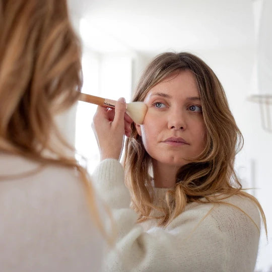 Woman applying makeup in front of a mirror using a Bachca powder brush with a wooden handle, gently sweeping blush or face powder onto her cheek.