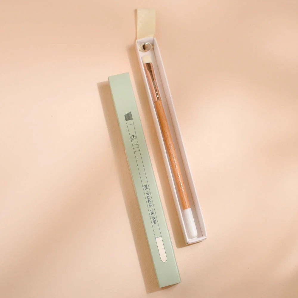 Woman applying eyeshadow with a Bachca brush featuring a wooden handle, concentrating on her upper eyelid makeup, in natural light.