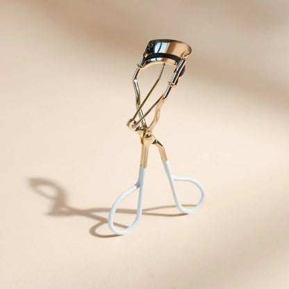 Bachca eyelash curler in gold metal with white handles, standing elegantly on a beige background.